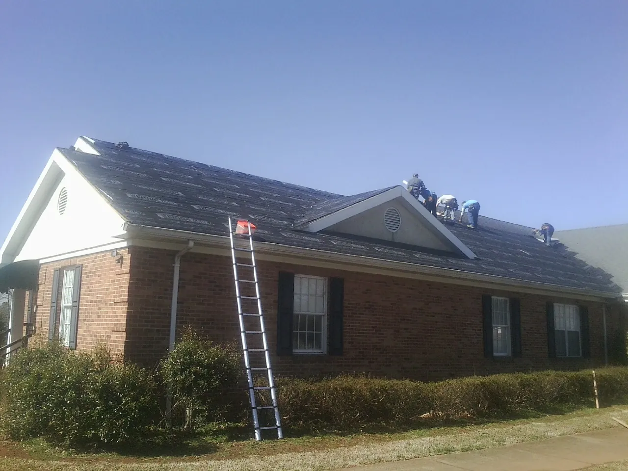 Crew tearing shingles off a brick ranch roof with a ladder set — Metal Roof Repair work by our crew in Martelle