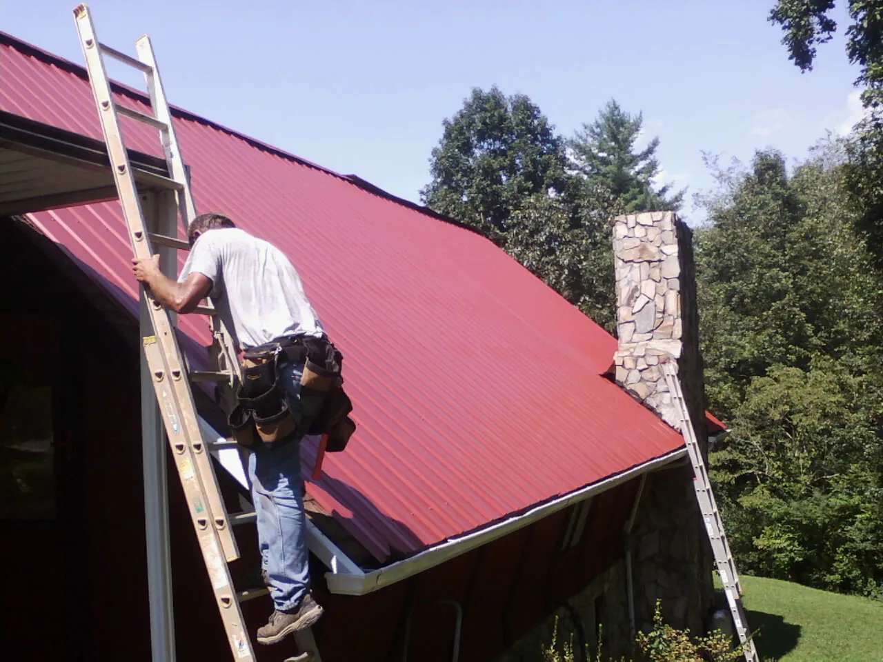 Roofer on a ladder beside a red metal roof and stone chimney — Soffit & Fascia Repair by Roofing Brothers in Martelle